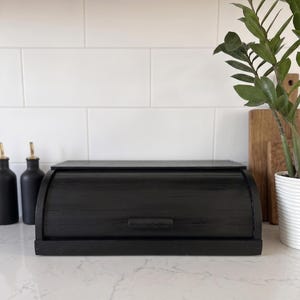 May include: A rectangular black wooden bread box with a curved roll-top lid and a small horizontal handle. The bread box features a smooth, dark finish and a solid base, designed for countertop storage of baked goods in a kitchen setting.