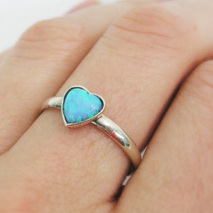 May include: A silver ring featuring a heart-shaped, light blue opal stone. The ring's band is smooth and polished. The opal has a shimmering, iridescent quality, with hints of green and blue. The ring is shown on a person's finger.