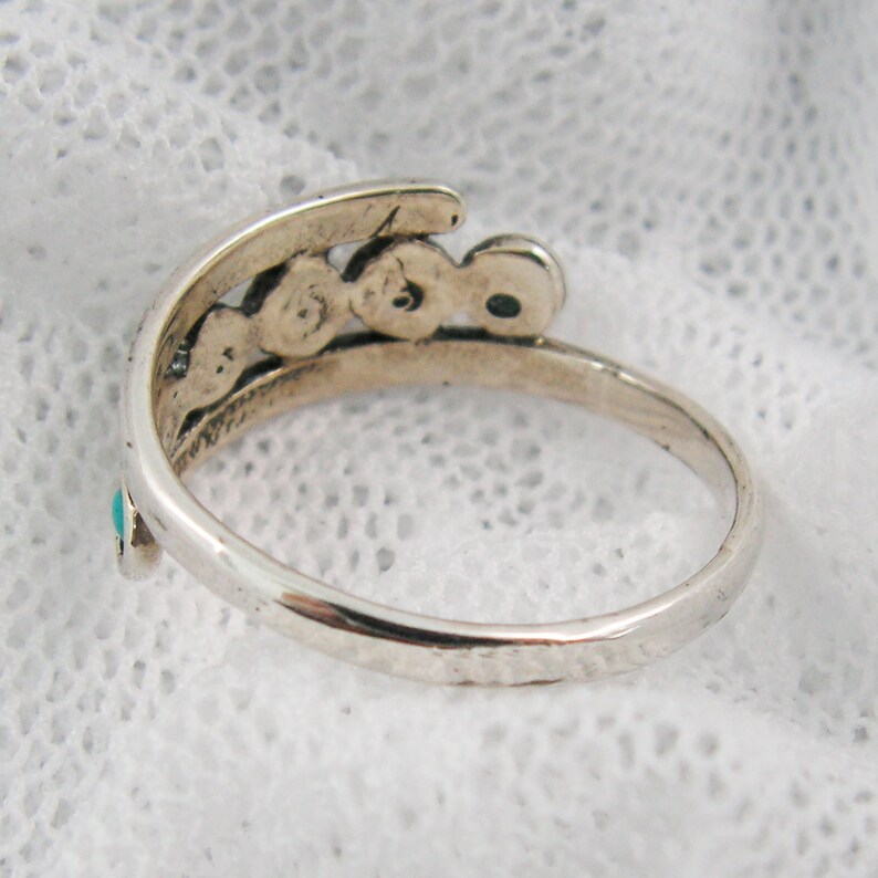 K&ouml;nnte beinhalten: Ein silberfarbener Ring mit geschwungenem Design. Der Ring hat kleine T&uuml;rkissteine und eine Reihe dekorativer kreisf&ouml;rmiger Elemente. Das Band hat eine geh&auml;mmerten Textur. Der Ring wird auf einer wei&szlig;en Spitzefl&auml;che gezeigt.