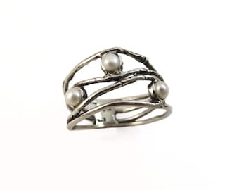 May include: A silver-toned ring with three round, white pearls. The ring has an open design with multiple bands and a textured, organic appearance. The pearls are evenly spaced across the top of the ring.