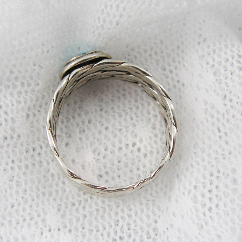 Puede incluir: Anillo plateado con un dise&ntilde;o de banda trenzada. El anillo presenta una peque&ntilde;a piedra redonda de color azul claro en la parte superior. El anillo est&aacute; sobre un fondo blanco texturizado.