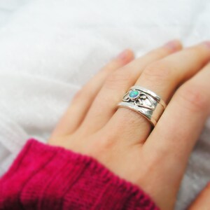 May include: A silver ring with an opal gemstone set in a delicate floral design. The ring is worn on a finger and the background is a white fabric.