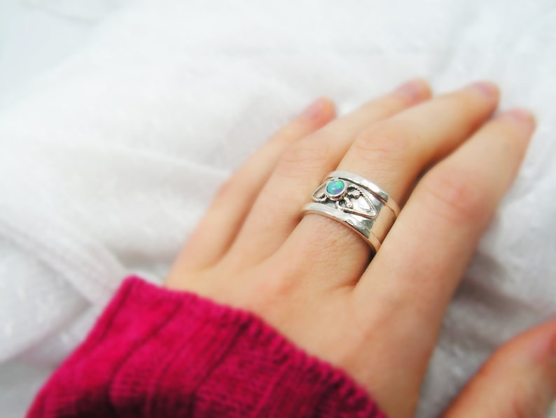 May include: A silver ring with an opal gemstone set in a delicate floral design. The ring is worn on a finger and the background is a white fabric.