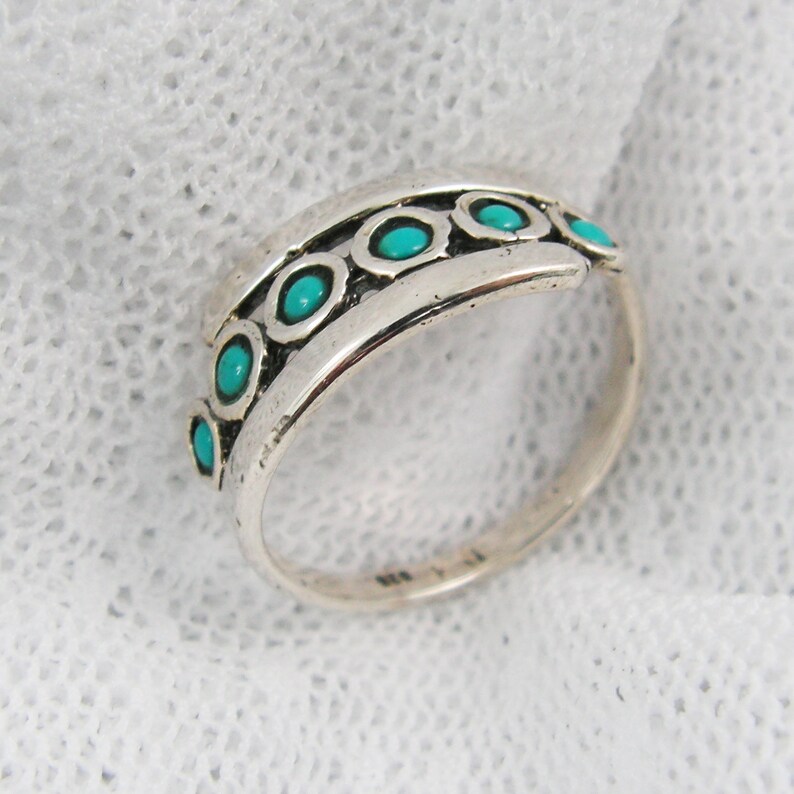 K&ouml;nnte beinhalten: Ein Silberring mit einem Band, das eine Reihe von t&uuml;rkisfarbenen Steinen in einzelnen Fassungen aufweist. Der Ring hat ein leicht gebogenes Design und eine polierte Oberfl&auml;che, die die leuchtend blau-gr&uuml;nen Steine zur Geltung bringt.