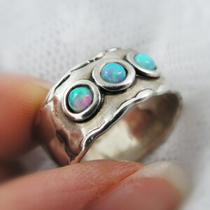 May include: A wide silver ring with three round, bezel-set opal stones. The opals display a play of color with hues of blue, green, and pink. The ring has a textured, organic design.
