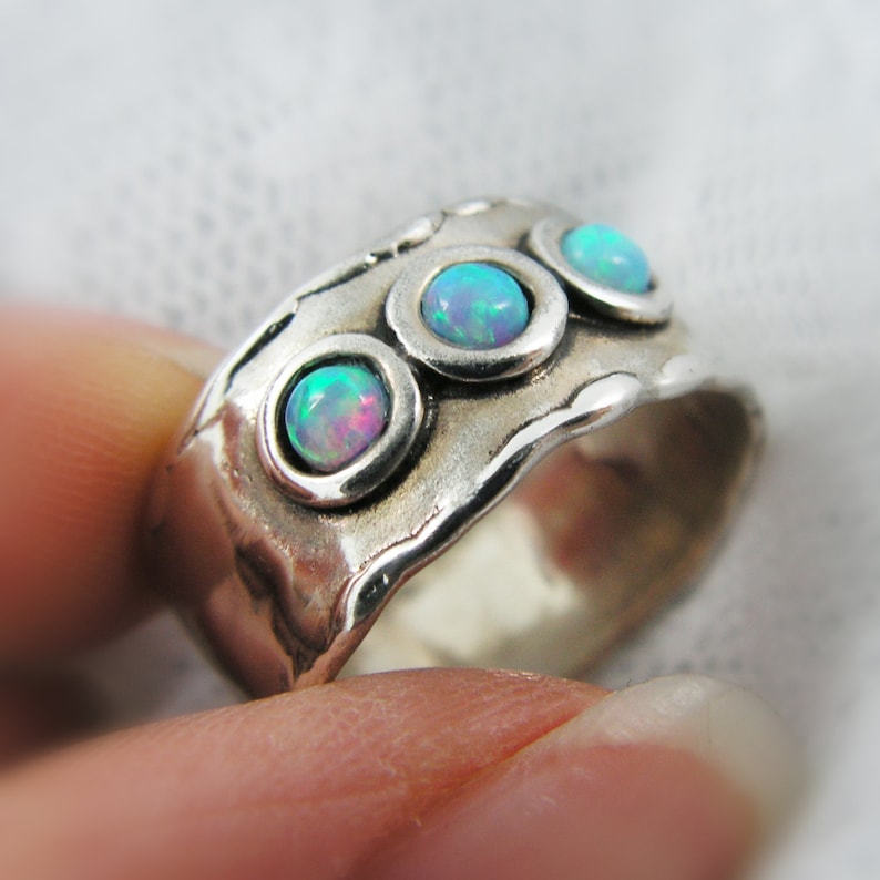 May include: A wide silver ring with three round, bezel-set opal stones. The opals display a play of color with hues of blue, green, and pink. The ring has a textured, organic design.
