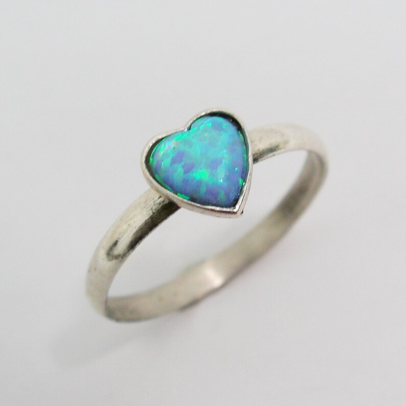 May include: A silver ring featuring a heart-shaped opal gemstone. The opal displays a vibrant mix of blue and green hues. The ring has a simple, elegant design, with a smooth, polished band.
