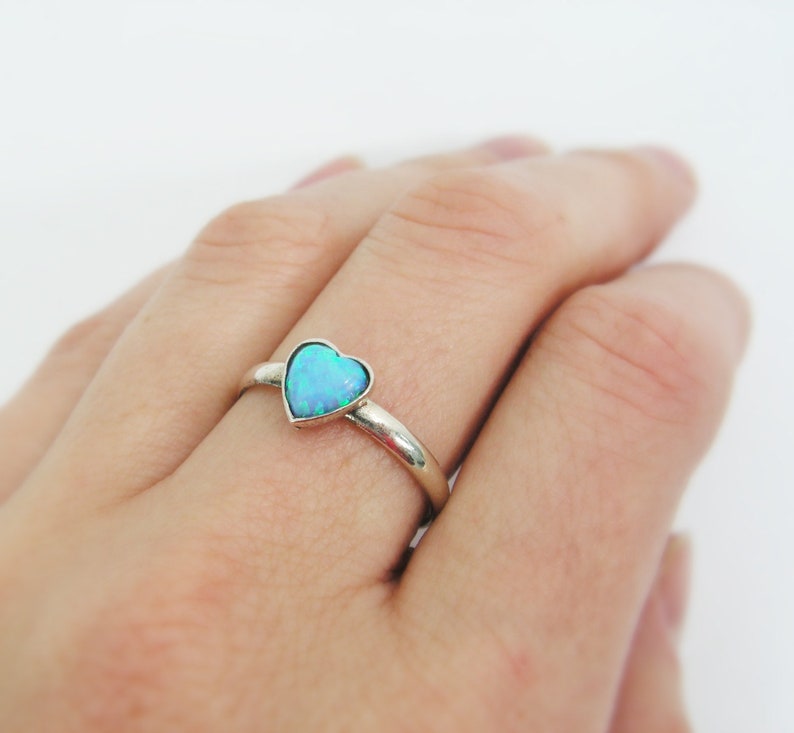 May include: A silver ring featuring a heart-shaped, light blue opal stone. The ring's band is smooth and polished. The opal has a shimmering, iridescent quality, with hints of green and blue. The ring is shown on a person's finger.