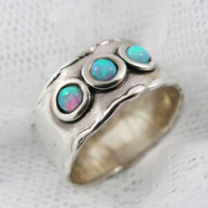 May include: A wide, silver-toned ring featuring three round, bezel-set opals. The opals display a mix of blue, green, and pink hues. The ring has a textured, hammered appearance with a wavy edge, giving it a handcrafted look.