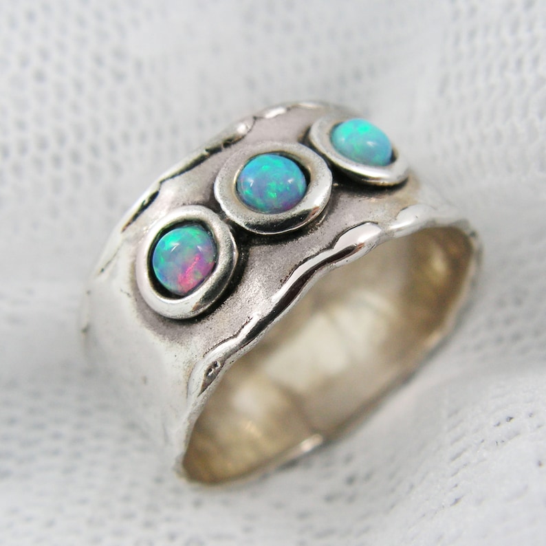 May include: A wide, silver-toned ring featuring three round, bezel-set opals. The opals display a mix of blue, green, and pink hues. The ring has a textured, hammered appearance with a wavy edge, giving it a handcrafted look.