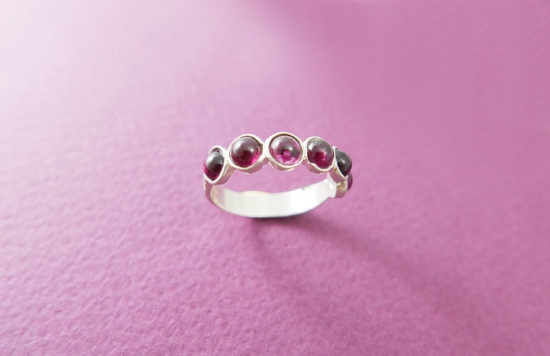 Pu&ograve; includere: Un anello d'argento con sei gemme rotonde di colore rosso scuro. L'anello &egrave; esposto su uno sfondo viola, evidenziando il design e il colore del gioiello.