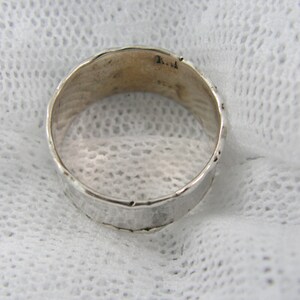 May include: A silver ring with a textured surface. The ring has a hammered appearance and a slightly uneven edge. The inside of the ring has some markings. The ring is set against a white, textured background.