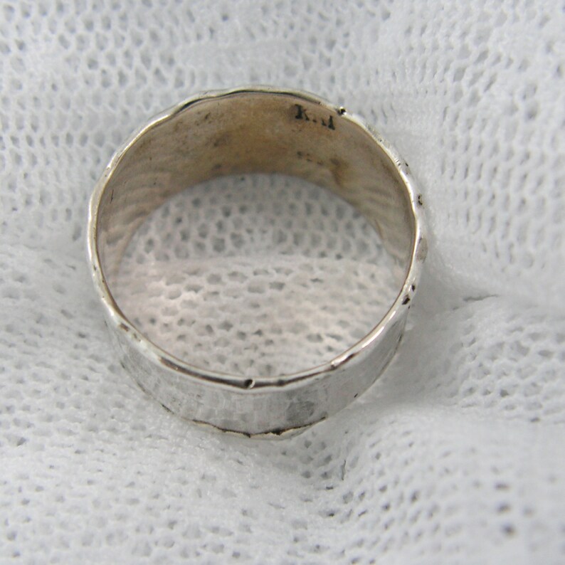 May include: A silver ring with a textured surface. The ring has a hammered appearance and a slightly uneven edge. The inside of the ring has some markings. The ring is set against a white, textured background.