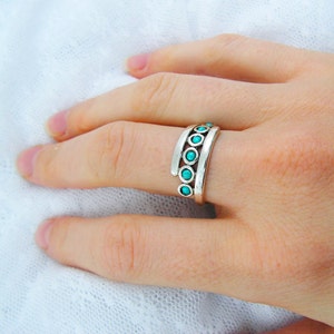 K&ouml;nnte beinhalten: Ein silberner Ring mit t&uuml;rkisfarbenen Steinen. Der Ring hat ein einzigartiges Design mit drei parallelen B&auml;ndern, die jeweils kleine, runde Steine halten. Der Ring wird an einem Finger getragen und zeigt seine komplizierten Details und die lebendige Farbe.