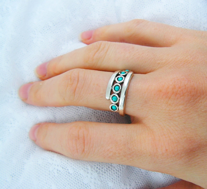 K&ouml;nnte beinhalten: Ein silberner Ring mit t&uuml;rkisfarbenen Steinen. Der Ring hat ein einzigartiges Design mit drei parallelen B&auml;ndern, die jeweils kleine, runde Steine halten. Der Ring wird an einem Finger getragen und zeigt seine komplizierten Details und die lebendige Farbe.