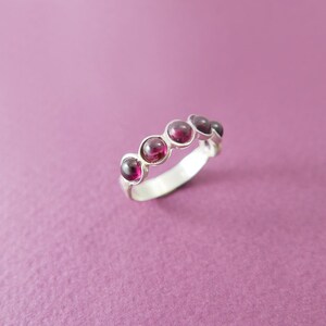Pu&ograve; includere: Un anello d'argento con cinque pietre preziose rotonde di colore rosso intenso, incastonate singolarmente. La fascia dell'anello &egrave; liscia e lucida, e le pietre sono equidistanti. L'anello &egrave; esposto su uno sfondo viola.