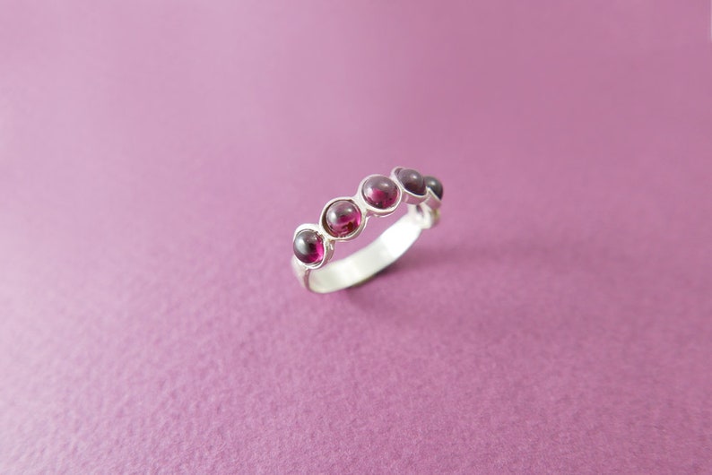 Pu&ograve; includere: Un anello d'argento con cinque pietre preziose rotonde di colore rosso intenso, incastonate singolarmente. La fascia dell'anello &egrave; liscia e lucida, e le pietre sono equidistanti. L'anello &egrave; esposto su uno sfondo viola.