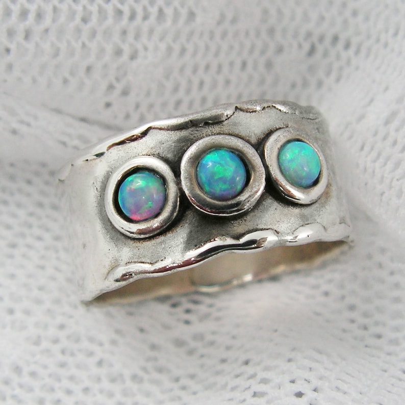May include: A wide silver ring with three bezel-set opal stones. The opals display a play of color with hues of blue, green, and pink. The ring has a textured, hammered appearance, giving it a rustic, handcrafted feel. The opals are round and set in circular bezels.