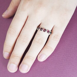 Pu&ograve; includere: Un anello d'argento con sei pietre preziose rotonde di colore rosso scuro. L'anello &egrave; su una mano, con uno sfondo viola.