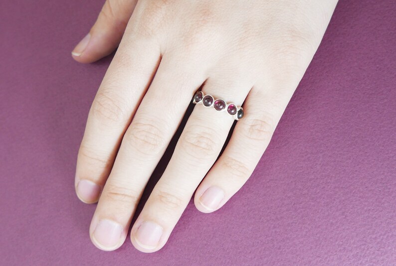Pu&ograve; includere: Un anello d'argento con sei pietre preziose rotonde di colore rosso scuro. L'anello &egrave; su una mano, con uno sfondo viola.