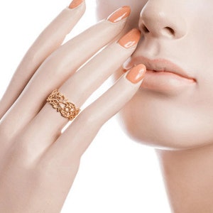 May include: A gold-colored ring with an openwork floral design. The ring is on a mannequin hand with orange-painted nails. The ring's intricate details are visible, showcasing a delicate and ornate style. The ring is a piece of jewelry.