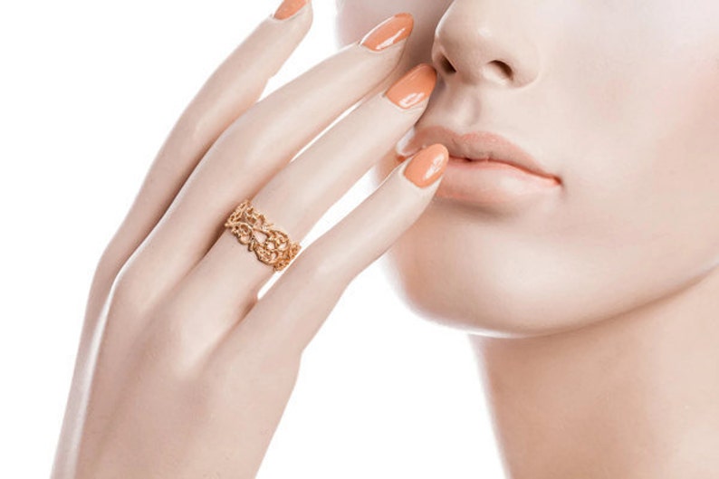May include: A gold-colored ring with an openwork floral design. The ring is on a mannequin hand with orange-painted nails. The ring's intricate details are visible, showcasing a delicate and ornate style. The ring is a piece of jewelry.