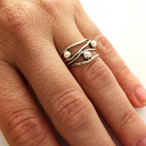 May include: A silver ring with a unique, organic design featuring three small, white pearls. The ring has a textured, branch-like appearance and is worn on a finger against a white background.