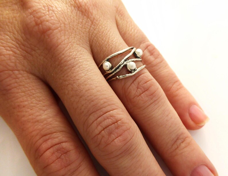 May include: A silver ring with a unique, organic design featuring three small, white pearls. The ring has a textured, branch-like appearance and is worn on a finger against a white background.