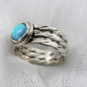 Puede incluir: Anillo de plata con un dise&ntilde;o de banda trenzada. El anillo presenta una piedra preciosa de &oacute;palo azul ovalada e iridiscente engastada en un bisel texturizado. La banda del anillo tiene una apariencia trenzada, agregando una textura &uacute;nica a la joya.