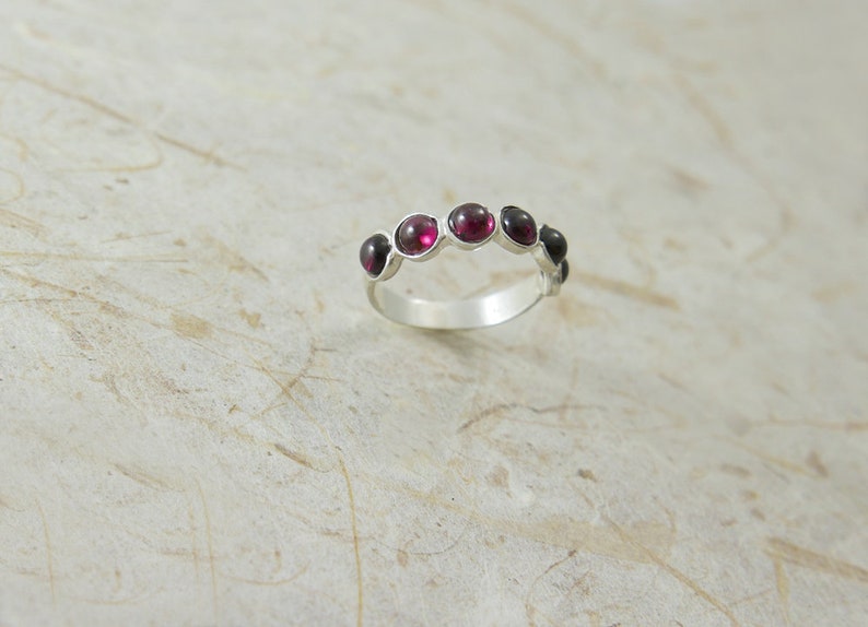 Pu&ograve; includere: Un anello d'argento con sette pietre preziose rotonde, di colore rosso intenso, incastonate lungo la fascia. Le pietre sono tenute in castoni individuali. L'anello &egrave; mostrato su una superficie strutturata bianco sporco.