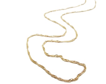 Puede incluir: Un delicado collar dorado con un diseño de cuerda trenzada. La cadena es fina y parece ser de metal. El collar se muestra sobre un fondo blanco, resaltando sus intrincados detalles y su elegante simplicidad.