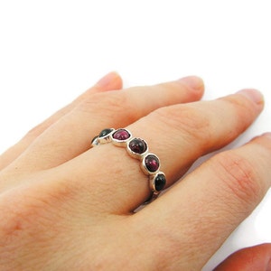 Pu&ograve; includere: Un anello d'argento con una serie di pietre preziose rotonde, rosso scuro e nere. L'anello &egrave; presentato su uno sfondo bianco, evidenziando il colore delle pietre preziose e il design dell'anello.