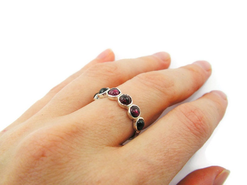 Pu&ograve; includere: Un anello d'argento con una serie di pietre preziose rotonde, rosso scuro e nere. L'anello &egrave; presentato su uno sfondo bianco, evidenziando il colore delle pietre preziose e il design dell'anello.