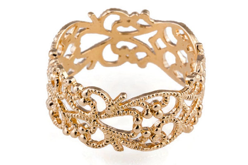 May include: A gold-colored ring with an intricate openwork design. The ring features a repeating pattern of floral and beaded elements, creating a delicate and ornate appearance. The ring is likely made of gold-toned metal.