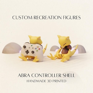 May include: Two yellow Abra figures, one holding a white game controller and one sitting, with text reading CUSTOM RECREATION FIGURES, ABRA CONTROLLER SHELL, and HANDMADE 3D PRINTED. The figures have pointed ears, brown chest markings, and tails.