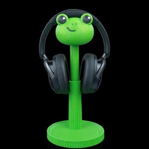 May include: A bright green frog-shaped headphone stand with a circular base and a vertical post. The frog head features large black eyes and a small smile. A pair of black over-ear headphones is resting on the frog head, supported by the stand.