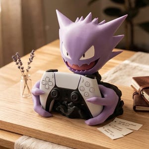 Puede incluir: Una figura del personaje Gengar en color morado que sirve como soporte para mando, sosteniendo un mando de videojuego blanco y negro. La figura tiene orejas puntiagudas, una amplia sonrisa y está montada sobre una base negra. Hay pequeñas tarjetas blancas con texto cerca.