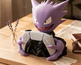 May include: A purple Gengar character figure acting as a controller stand, holding a white and black video game controller. The figure has pointed ears, a wide grin, and is mounted on a black base. Small white cards with text are placed nearby.