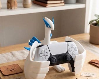 May include: A white and blue Lugia figure acting as a controller stand, holding a white and black game controller. The figure features large wings that cradle the controller. Small paper tags on the surface read Handmade 3D Print, Lugia Collector Item, and Functional Art.