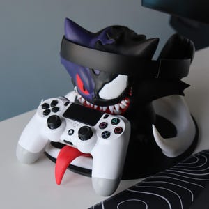 May include: A dual-purpose gaming accessory stand featuring a split-design character head with one purple side and one black side. The stand holds a black headset across the top and a white wireless game controller resting on a red tongue protrusion.