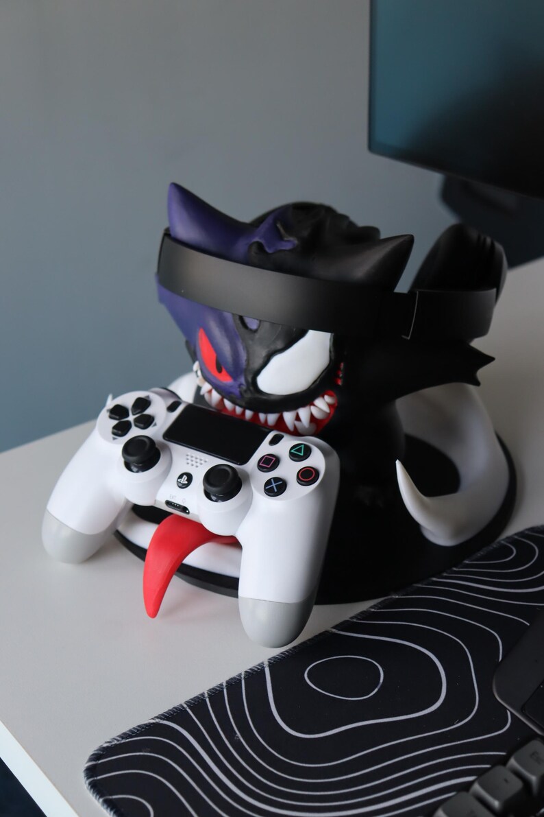 May include: A dual-purpose gaming accessory stand featuring a split-design character head with one purple side and one black side. The stand holds a black headset across the top and a white wireless game controller resting on a red tongue protrusion.