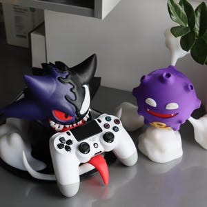 May include: A purple and black character figure with sharp teeth and red eyes holding a white game controller with a long red tongue. Beside it stands a smaller purple spherical character with white eyes and a yellow ring, surrounded by white smoke.