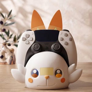May include: A white and black video game controller resting in a white character-shaped stand. The stand features orange ears, round orange cheeks, blue and yellow eyes, and a small yellow rectangular patch on the forehead area.