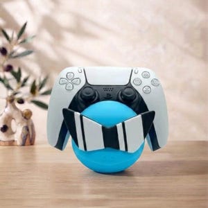 May include: A white and black video game controller resting on a bright blue spherical stand. The stand features a decorative black and white striped bowtie-shaped attachment on the front, positioned directly below the controller's central buttons.