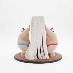 May include: A bookend set featuring two beige figures with dark hair, one wearing a blue loincloth and the other a red loincloth, positioned on a circular brown base. The figures are shown pushing against a stack of white paper pages.