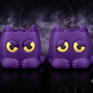 May include: Two purple cat-shaped containers with pointed ears and large yellow eyes. Each figure features a rounded body, small front paws, and a hollow top opening. The design includes expressive, angled yellow eyes set into the solid purple structure.