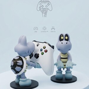 May include: Two light blue turtle-like figures on black circular bases. One figure holds a white game controller with black, green, red, and yellow buttons. Above them, a line drawing shows a skeletal hand holding a controller with icons labeled grip, universal fit, and desk art.