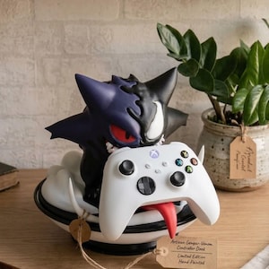 May include: A hand-painted controller dock featuring a hybrid Gengar and Venom character figure in purple and black, holding a white Xbox wireless controller. The base is a black and white bowl shape with a tag reading Artiean Gengar-Venom Controller Dock.