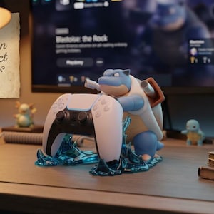 May include: A blue and beige Blastoise figurine acting as a controller stand for a white and black game controller, featuring translucent blue water-effect base details and a paper tag reading Artisan Product Hand-Painted in the upper left corner.