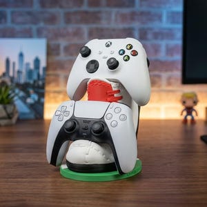 May include: A white and red sneaker-shaped controller stand holding a white Xbox wireless controller on top and a white and black PlayStation DualSense controller in the front, all resting on a circular green base with a flat surface.